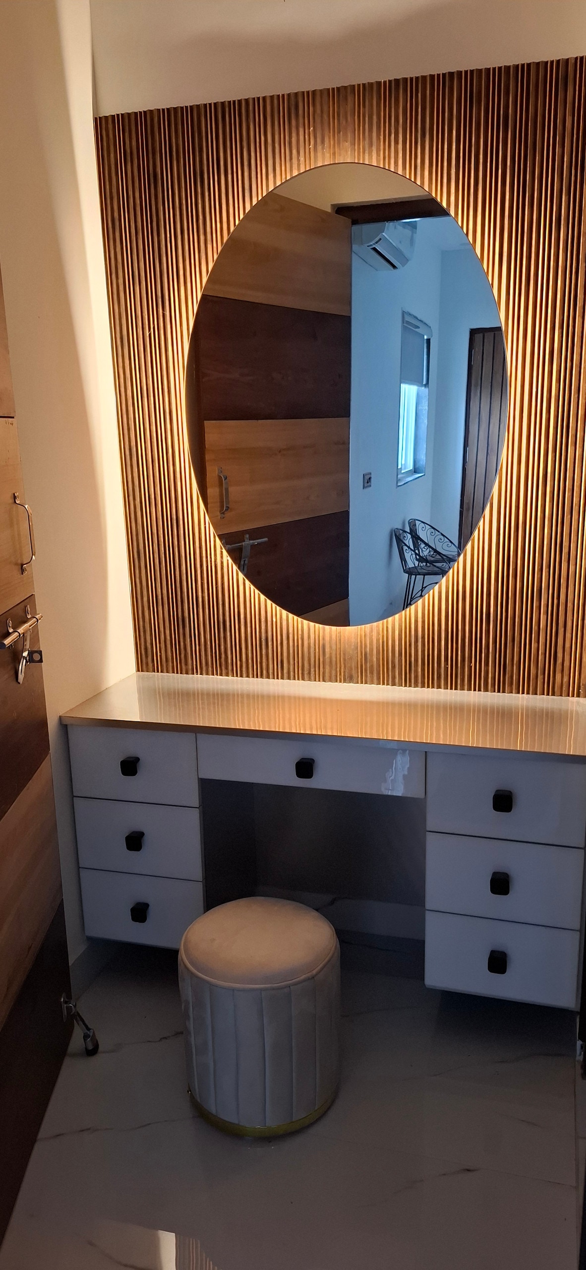 A stylish vanity area is showcased with a large oval mirror, framed by wooden panels. The setup includes a sleek countertop with multiple drawers. A cushioned stool is positioned in front, complementing the modern aesthetics of the space.