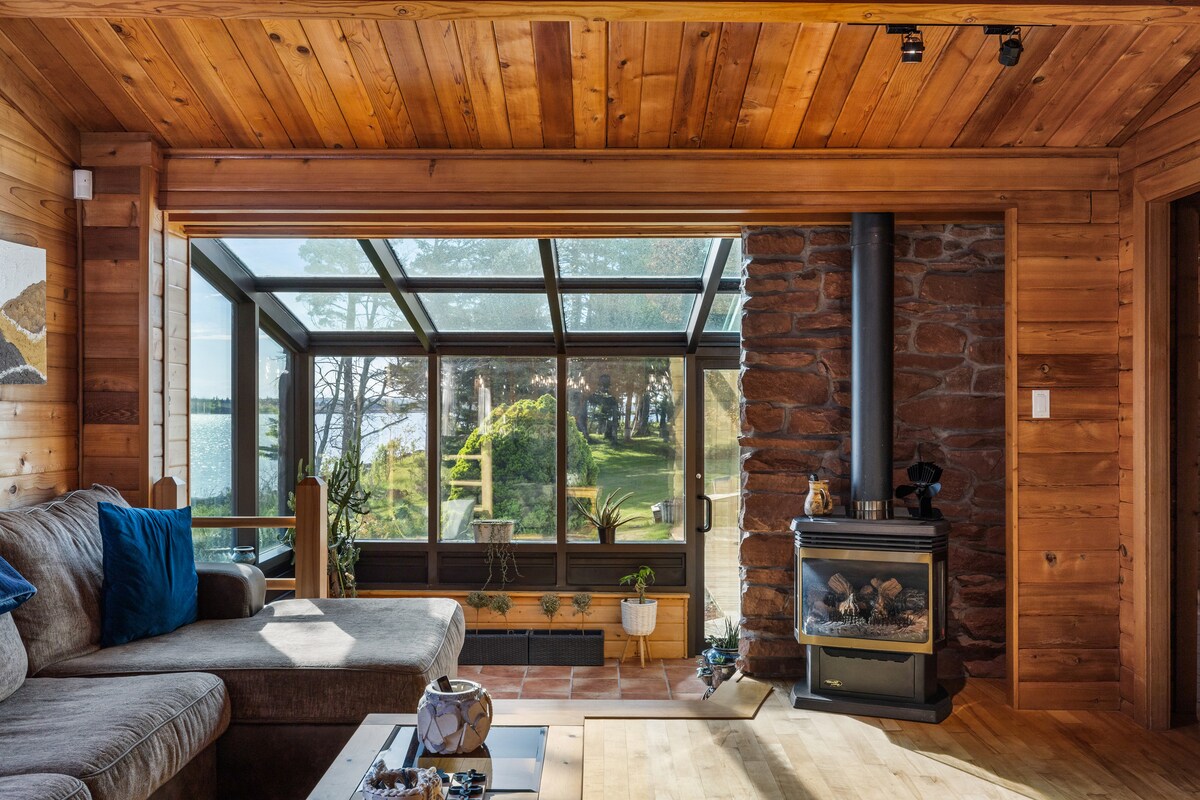 A cozy living area features warm wooden paneling and a comfortable sofa, complemented by a wooden coffee table. A stone fireplace provides warmth, while large glass windows allow natural light and views of the yard outside, creating a connection with the serene nature beyond.