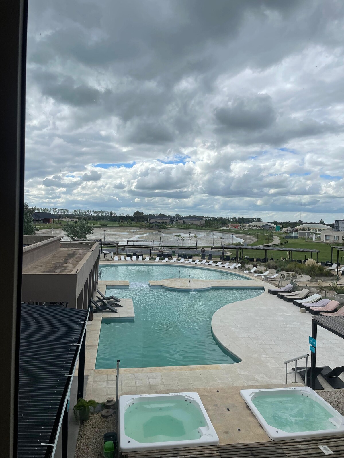 A spacious pool with gentle curves is surrounded by lounge chairs and natural landscapes. Two jacuzzis are visible nearby, offering a relaxing option. The sky is filled with clouds, casting soft shadows on the pool area, enhancing the serene atmosphere.