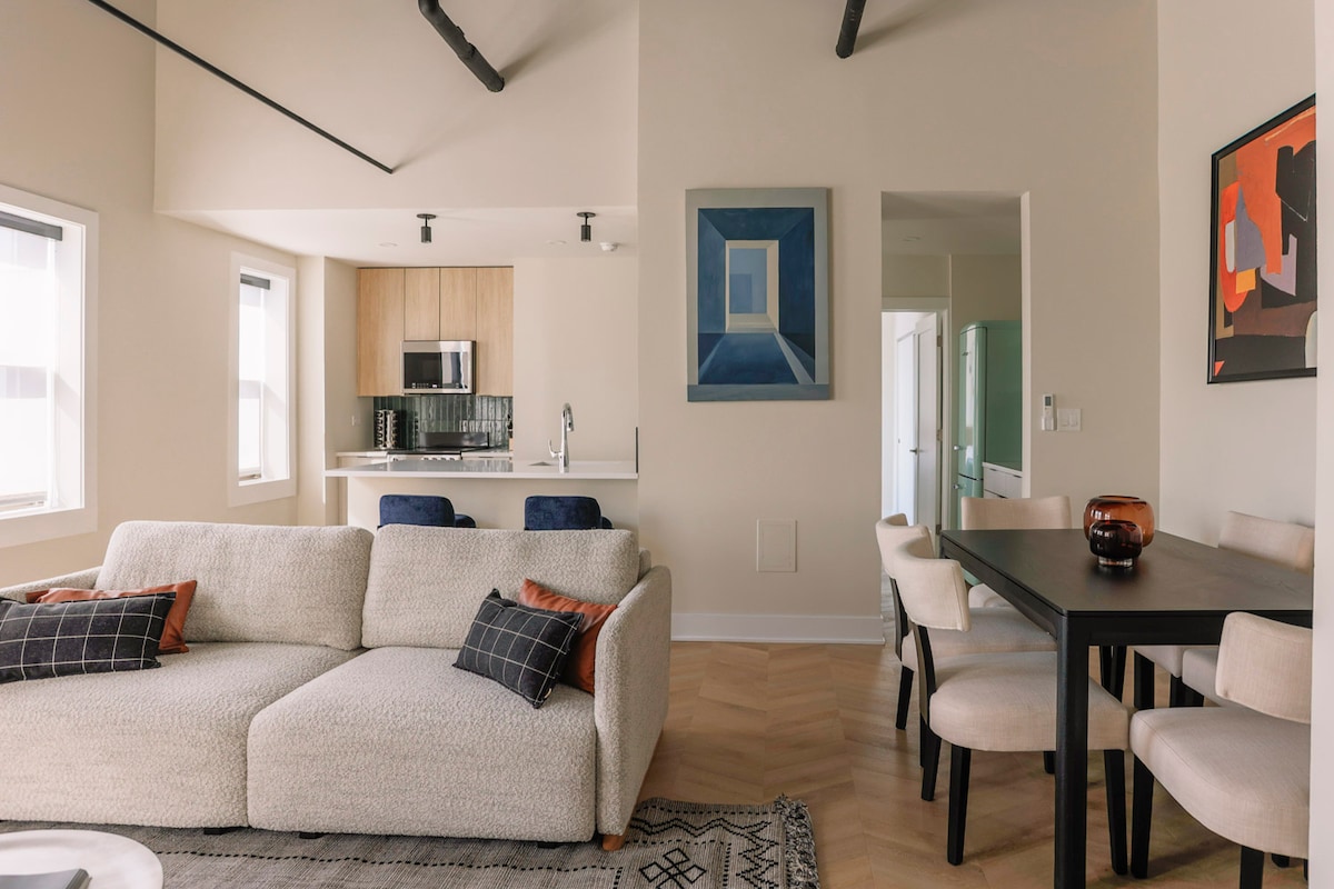The spacious living and dining area features mid-century modern furnishings. A large light-colored sofa faces the dining table, which seats four, while a minimalist kitchen is visible in the background, showcasing wooden cabinetry and high-end appliances. Natural light enters through large windows, brightening the space.