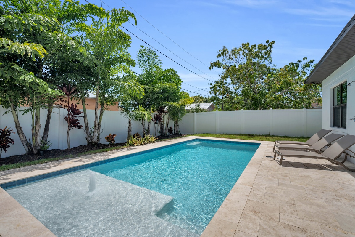 A serene backyard features a private, heated saltwater pool surrounded by lush greenery. Sun loungers are placed nearby on a tiled patio, providing a perfect spot for relaxation. The bright blue sky and clear water create an inviting outdoor atmosphere.