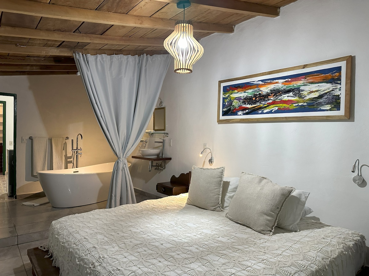 A spacious sleeping area features a large bed dressed with light neutral linens and decorative pillows. A contemporary pendant light hangs above, while a colorful piece of art adorns the wall. Nearby, a freestanding bathtub is partially visible, separated by a flowing curtain.