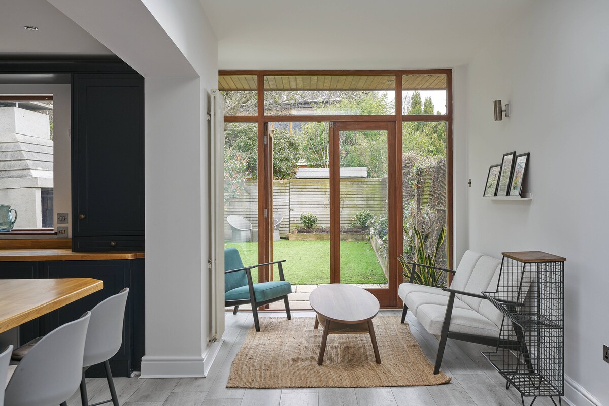 A light-filled living area features large glass doors opening to a well-maintained garden. Comfortable seating is arranged around a round table on a natural fiber rug. One chair is upholstered in teal, and a streamlined black side table is positioned nearby.