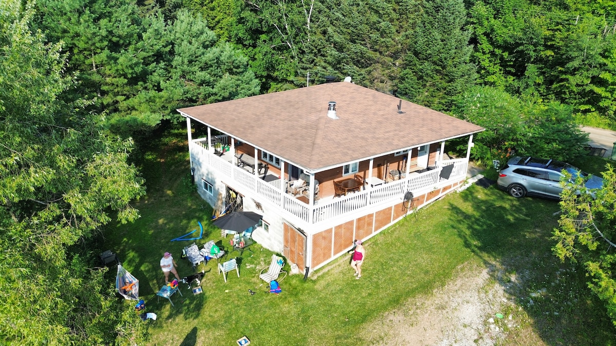 The chalet is captured from an aerial view, showcasing a spacious deck surrounded by greenery. A well-maintained yard is visible, featuring areas for relaxation and outdoor activities. Nearby, vehicles are parked, indicating accessibility and convenience.