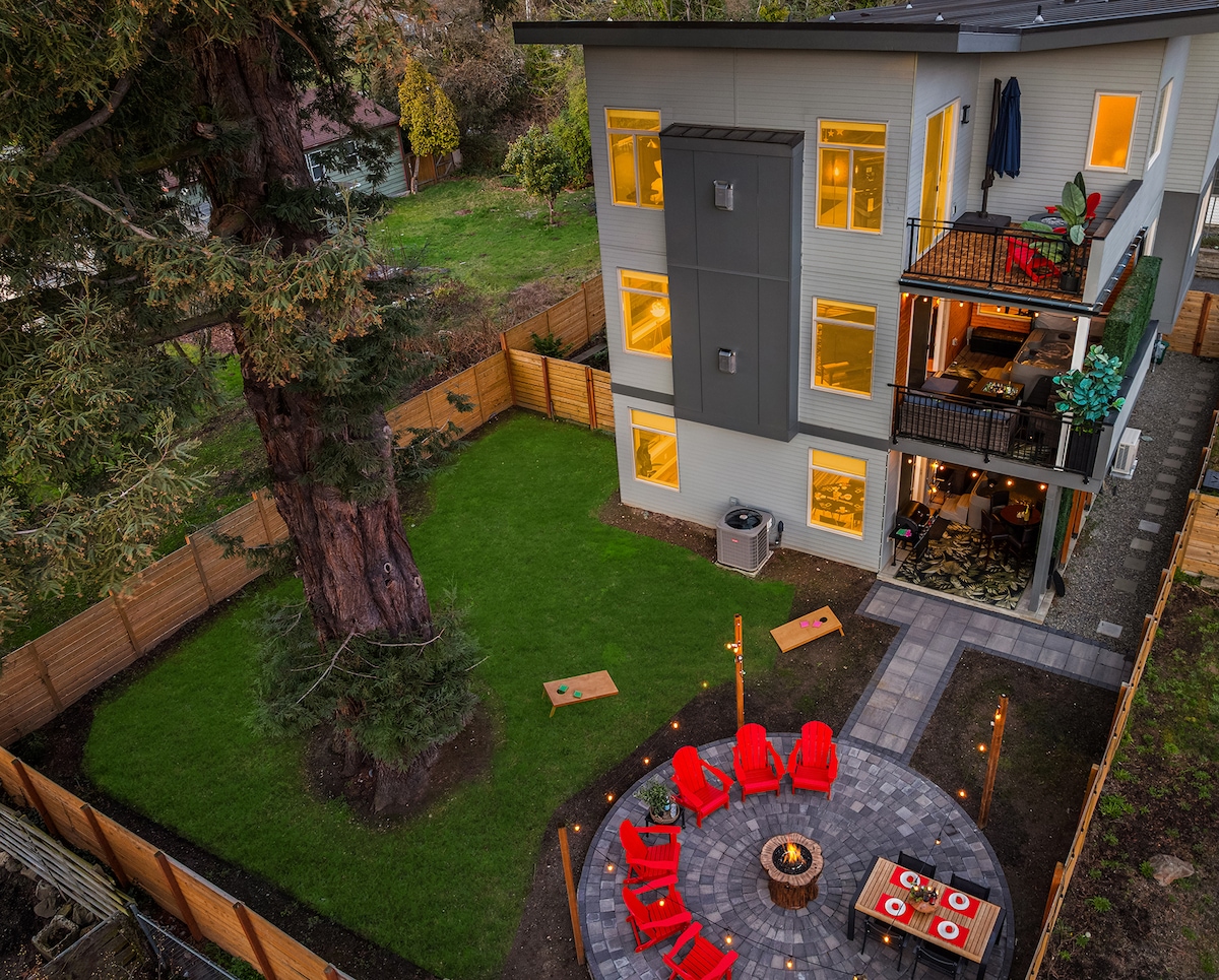 The exterior showcases a three-story home framed by a spacious green lawn and a large tree. A circular patio with bright red chairs surrounds a firepit, with cornhole boards on the grass. Warm light emanates from the windows, creating a welcoming ambiance.