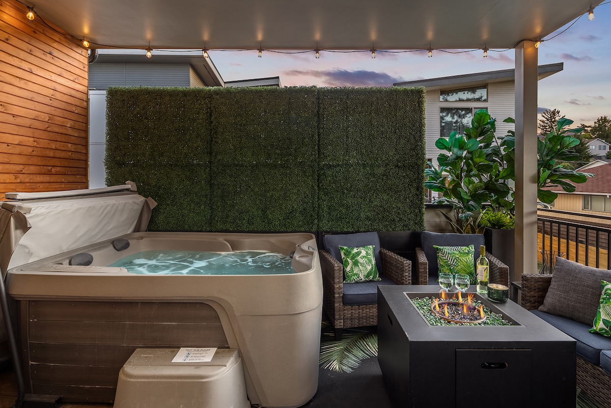 A private outdoor space is highlighted by a hot tub surrounded by lush greenery. Comfortable seating arrangements are visible, complemented by decorative pillows. A cozy firepit is positioned at the center, creating an inviting atmosphere for relaxation in the evening light.