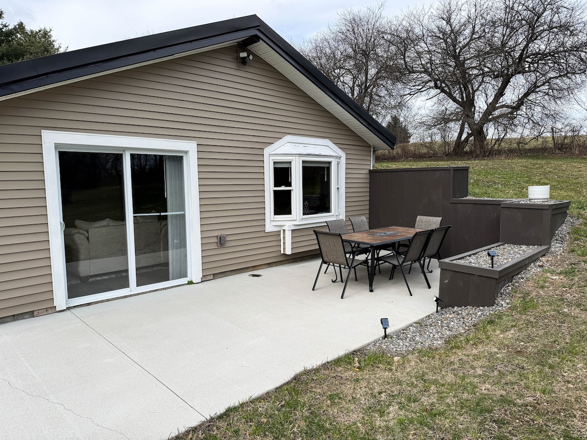 An outdoor patio area features a table surrounded by six chairs and is enclosed by low stone walls. The sliding glass doors lead into the house, while a grassy yard extends beyond the patio, offering a serene view of tree-covered hills.