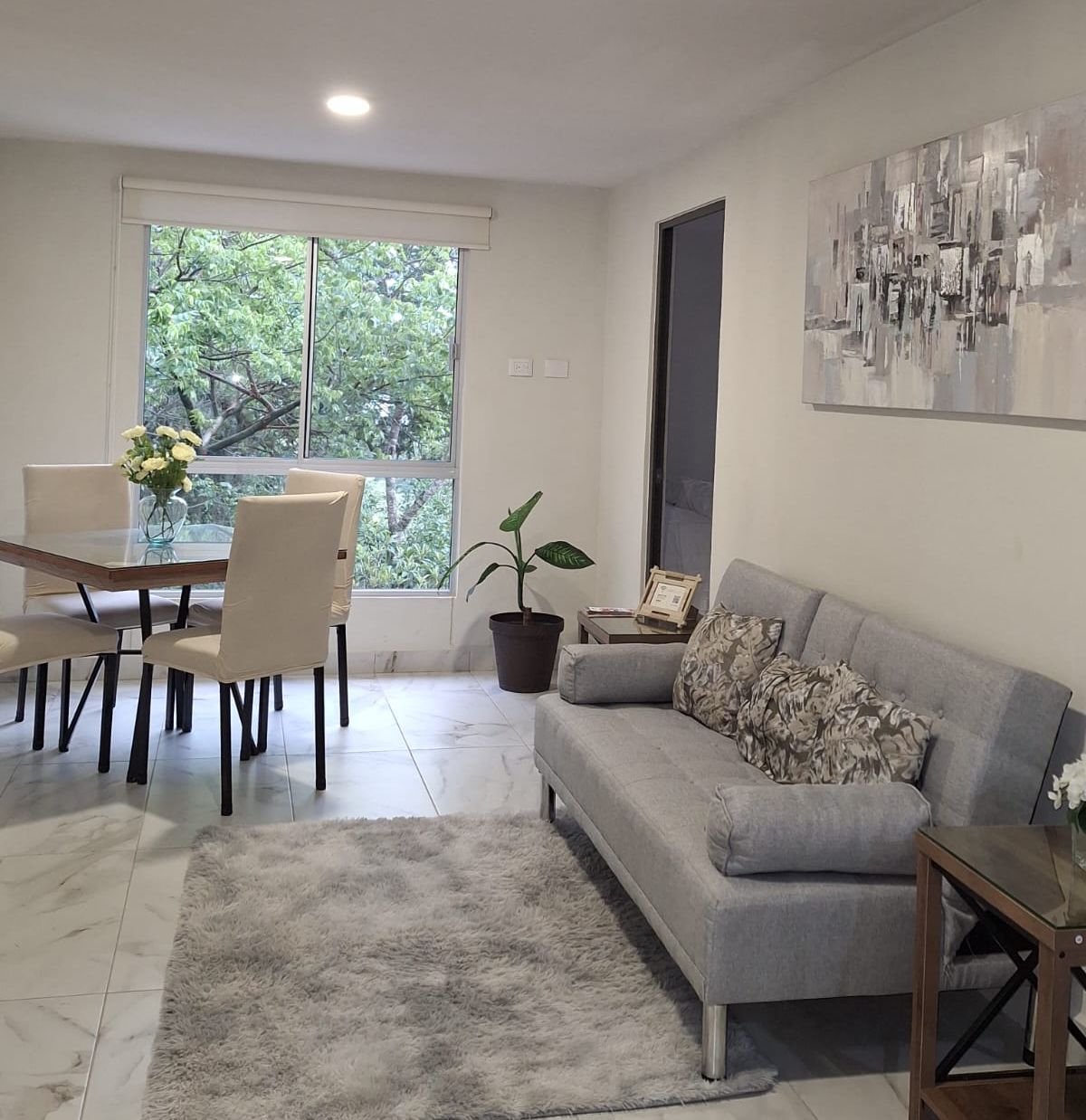 A modern living area features a comfortable gray sofa adorned with patterned pillows. A dining table with beige chairs is positioned near a large window, allowing natural light to illuminate the space. A potted plant adds a touch of greenery to the cozy environment.