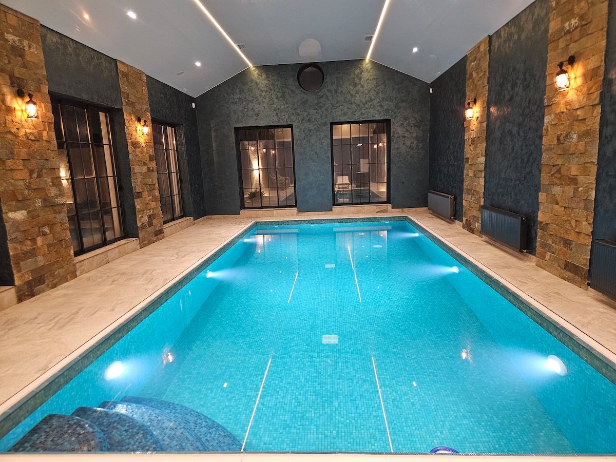 An indoor swimming pool is surrounded by textured stone walls and large glass windows. The water is a clear turquoise, reflecting the soft lighting from above. Smooth tiles cover the pool area, creating a serene space for relaxation and recreation.