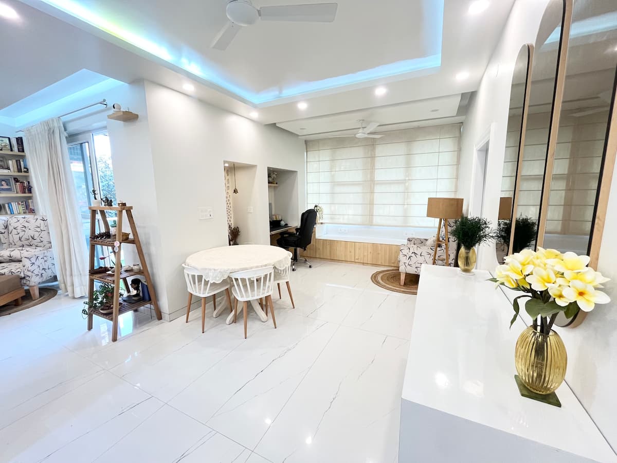 A bright and airy living space is highlighted by a round dining table surrounded by six chairs. A cozy seating area with two armchairs is visible to the side. Natural light pours in through large windows, and modern design elements add to the room's contemporary feel.