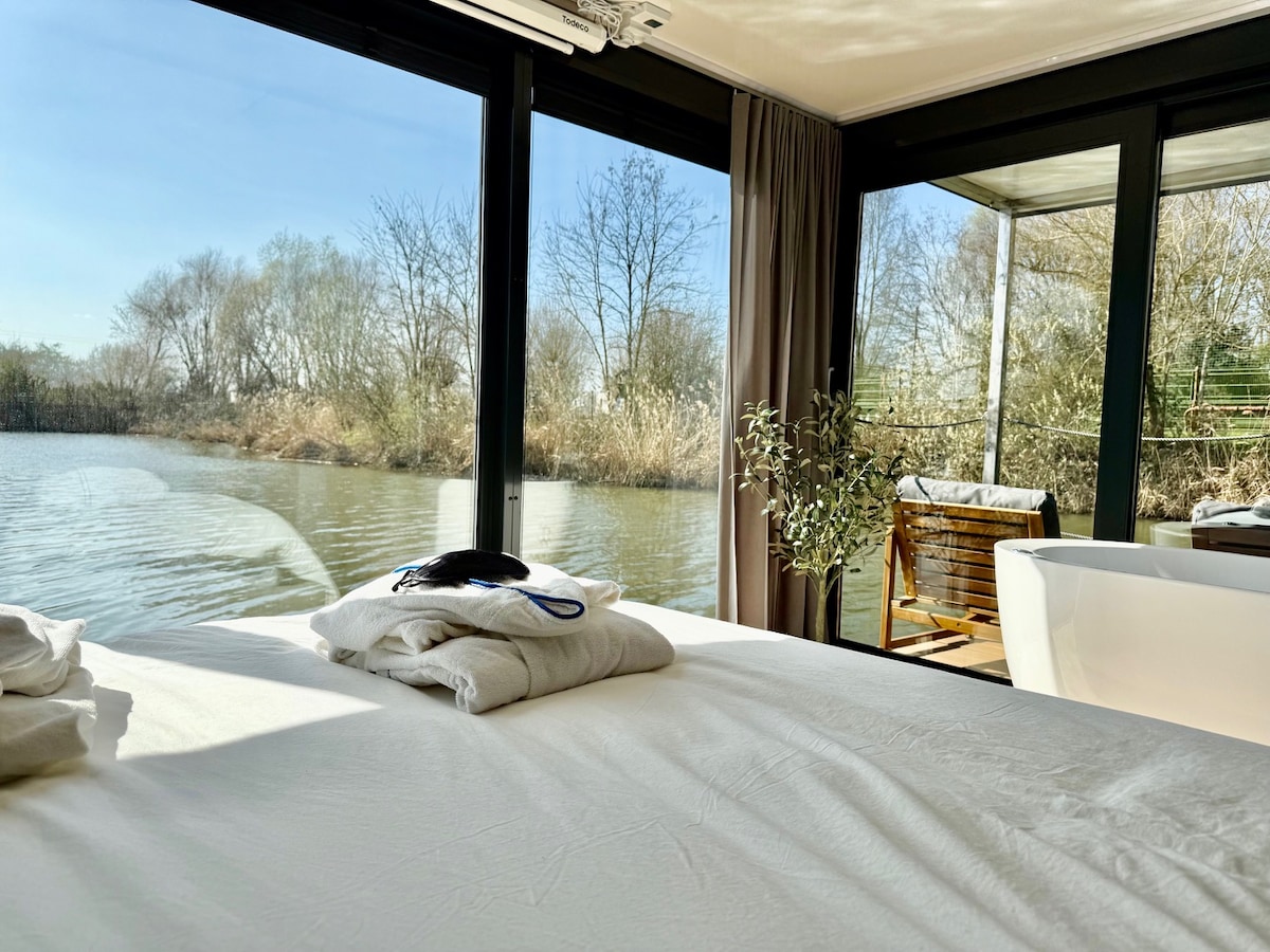 A spacious bed with neatly arranged linens is positioned near large glass windows, offering views of a calm waterway and surrounding trees. A comfortable chair sits on a deck outside, while a modern bathtub is visible in the corner, enhancing the relaxing environment.
