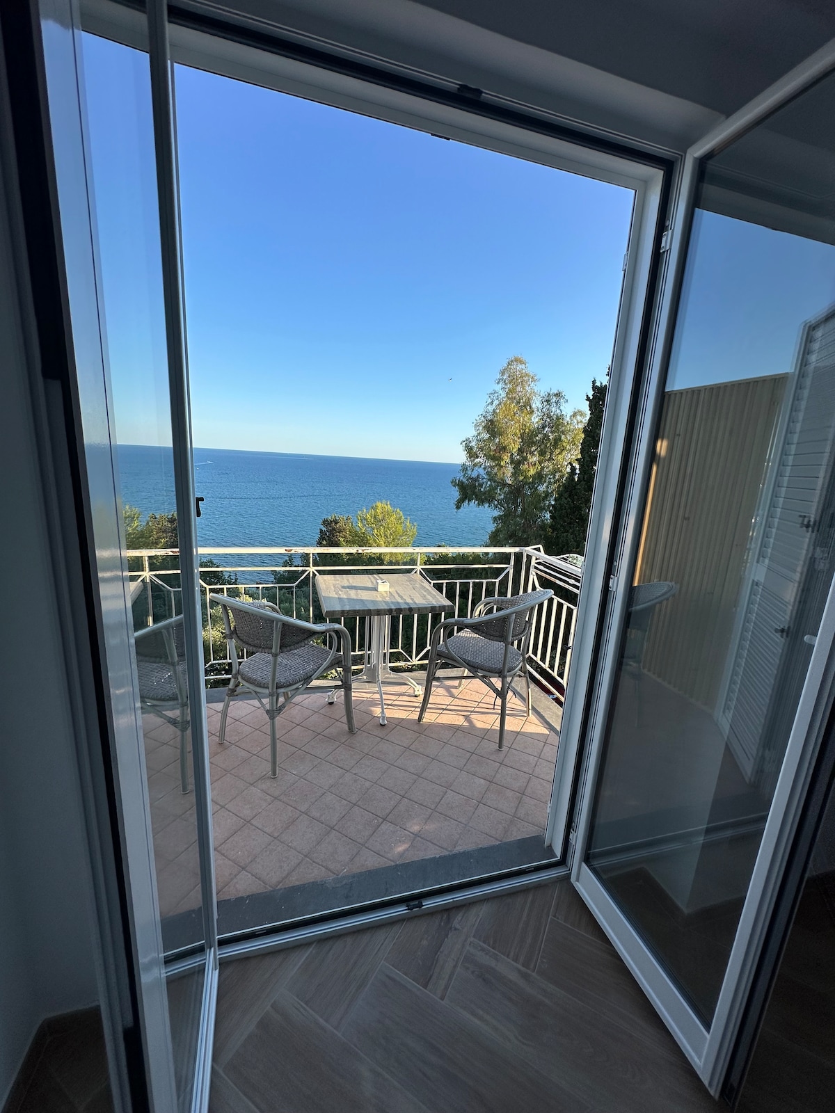 A pair of glass doors opens to a private terrace, showcasing a breathtaking view of the sea. Outdoor furniture, including a table and two chairs, is positioned on the tiled balcony, allowing for relaxation and dining while enjoying the scenic backdrop.