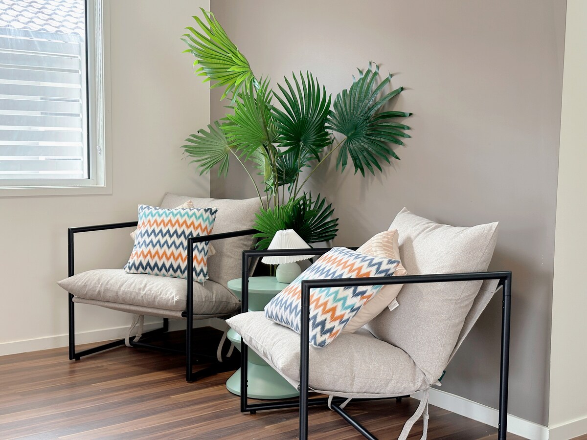 A comfortable sitting area features two light-colored armchairs, each adorned with patterned cushions. A small round table is positioned between them, accompanied by a lamp. Lush green plants provide a touch of nature against the neutral wall.