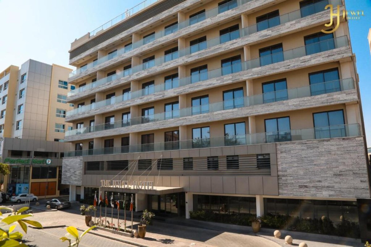 The exterior of a modern multi-story building is displayed, featuring a sleek façade with large glass windows. The entrance is marked by an awning and potted plants are positioned along the walkway. Nearby, an adjacent building is visible, contributing to the urban environment.