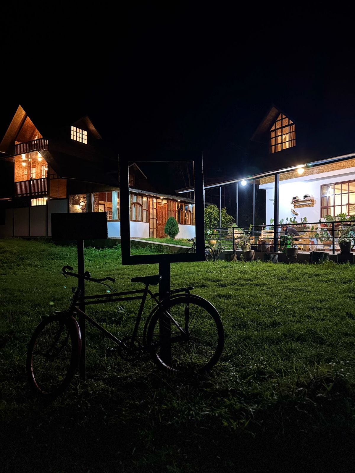 The image features a dimly lit exterior of a lodge at night, showcasing a charming building with triangular roofs and illuminated windows. A bicycle rests nearby on the green grass, and plants are visible on the porch, enhancing the connection to nature.