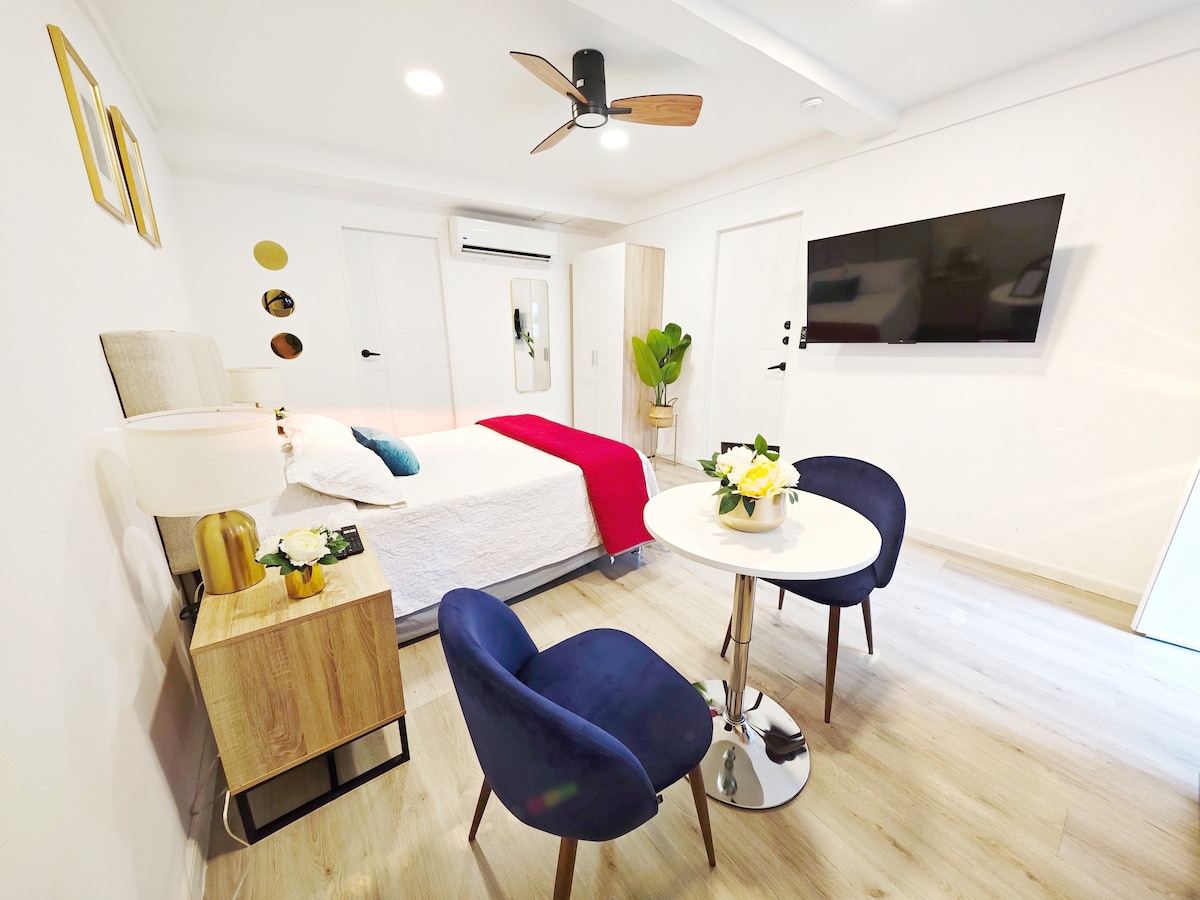 A bright and airy room features a spacious bed with a white quilt and decorative pillows. A small dining table with two blue chairs is positioned nearby. The room includes a flat-screen TV mounted on the wall, a modern ceiling fan, and a potted plant, adding a touch of greenery.