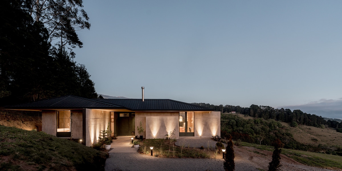 The exterior of a modern house is illuminated subtly by outdoor lighting, highlighting the minimalist architectural design. A gravel pathway leads to the entrance, surrounded by landscaped greenery and rolling hills in the background, creating a serene and peaceful setting.