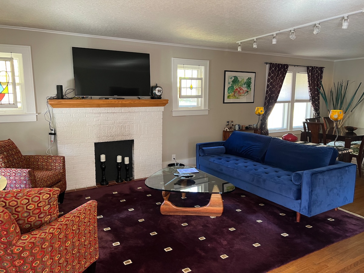 The living room features a bold blue sofa and two patterned chairs set on a deep purple carpet. A large television is mounted above a white brick fireplace, while a glass coffee table centers the seating area. Natural light is filtered through window treatments.