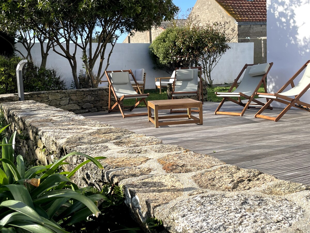 An outdoor terrace is highlighted by a wooden table and several reclining chairs. A manicured garden is present in the background, with lush green plants and trees providing a serene environment. A stone wall borders the terrace, offering a natural aesthetic.