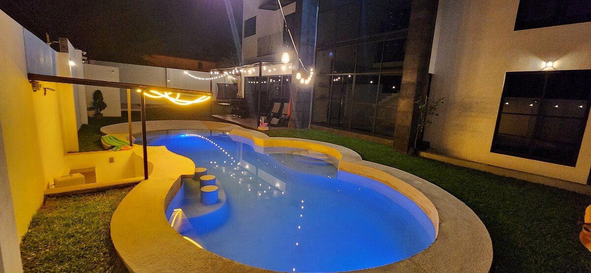 A well-lit swimming pool featuring a unique shape, surrounded by a lush green lawn. The pool includes seating areas and is illuminated with ambient lighting, enhancing the evening atmosphere. The backdrop showcases modern architecture with large windows, creating an inviting outdoor space.
