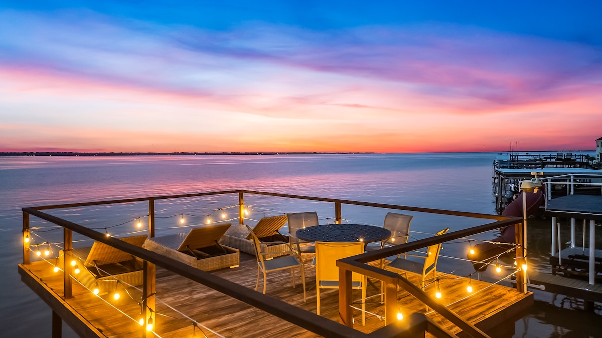 A spacious outdoor deck extends over the water, featuring a circular table surrounded by four chairs. Warm lights create a soft glow against the backdrop of a vibrant sunset, casting colorful reflections on the lake's surface.