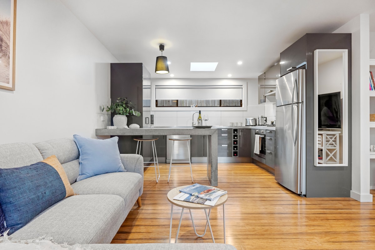 A well-equipped kitchen is visible, featuring modern appliances including a refrigerator and oven. A comfortable seating area is nearby, with a light-colored sofa adorned with a blue pillow. Natural light enters from a skylight, enhancing the warm wood flooring.