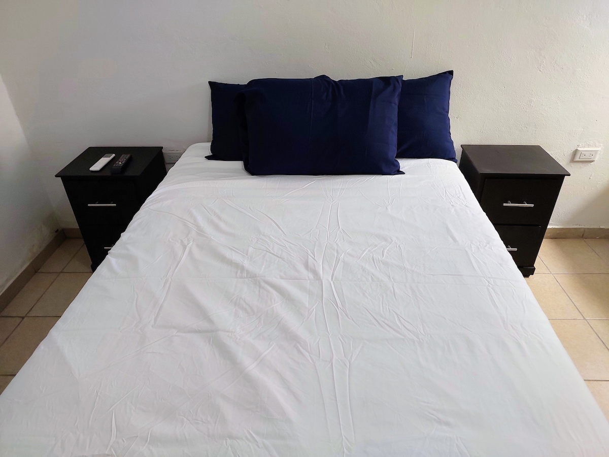 A cozy sleeping area features a spacious bed dressed in crisp white linens, complemented by dark blue pillows. Two nightstands with drawers are positioned on either side of the bed, providing additional surface space. Light-colored tiles cover the floor, enhancing the room's brightness.