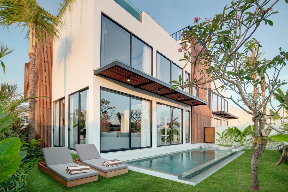 The modern villa exterior features large glass doors framed by wooden accents, providing a seamless transition to the outdoors. Lounge chairs are positioned beside a private pool, surrounded by lush greenery and trees, creating a serene outdoor space.