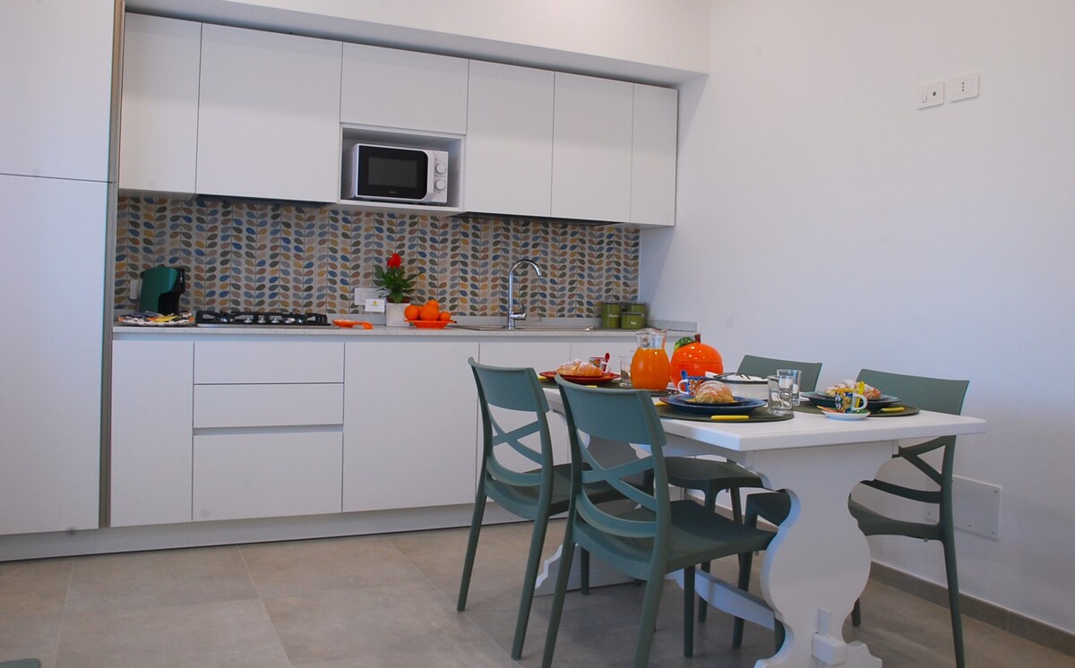 A modern kitchen area is furnished with white cabinetry and a tiled backsplash in earthy tones. A dining table for four is set with plates and seasonal decorations. A microwave and gas stove are present, contributing to the functional space.