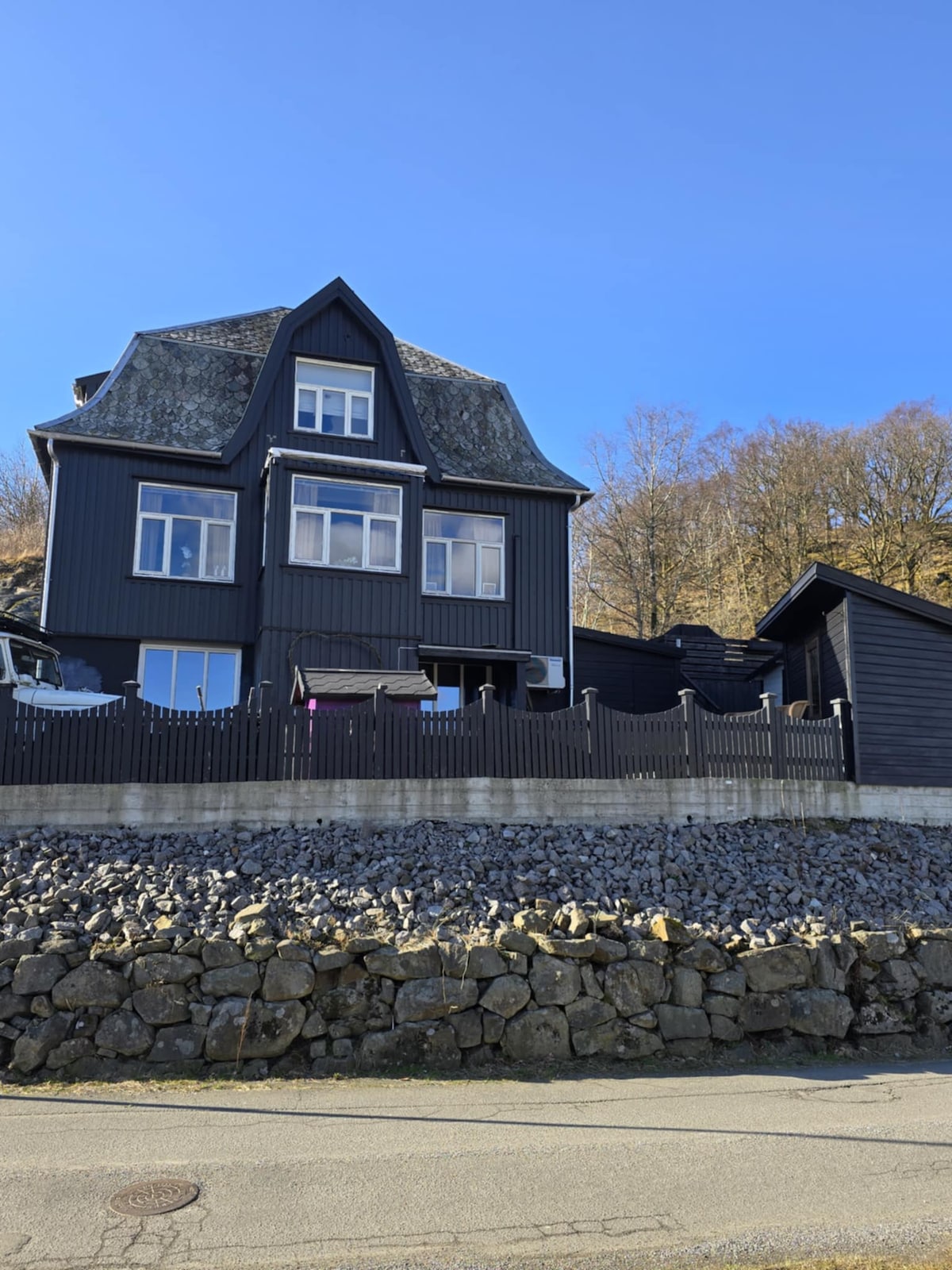 A charming black house features multiple levels and large windows, allowing natural light to fill the interior. The surrounding area includes stone landscaping and a low stone wall. A smaller black structure is visible nearby, complementing the main residence's design.