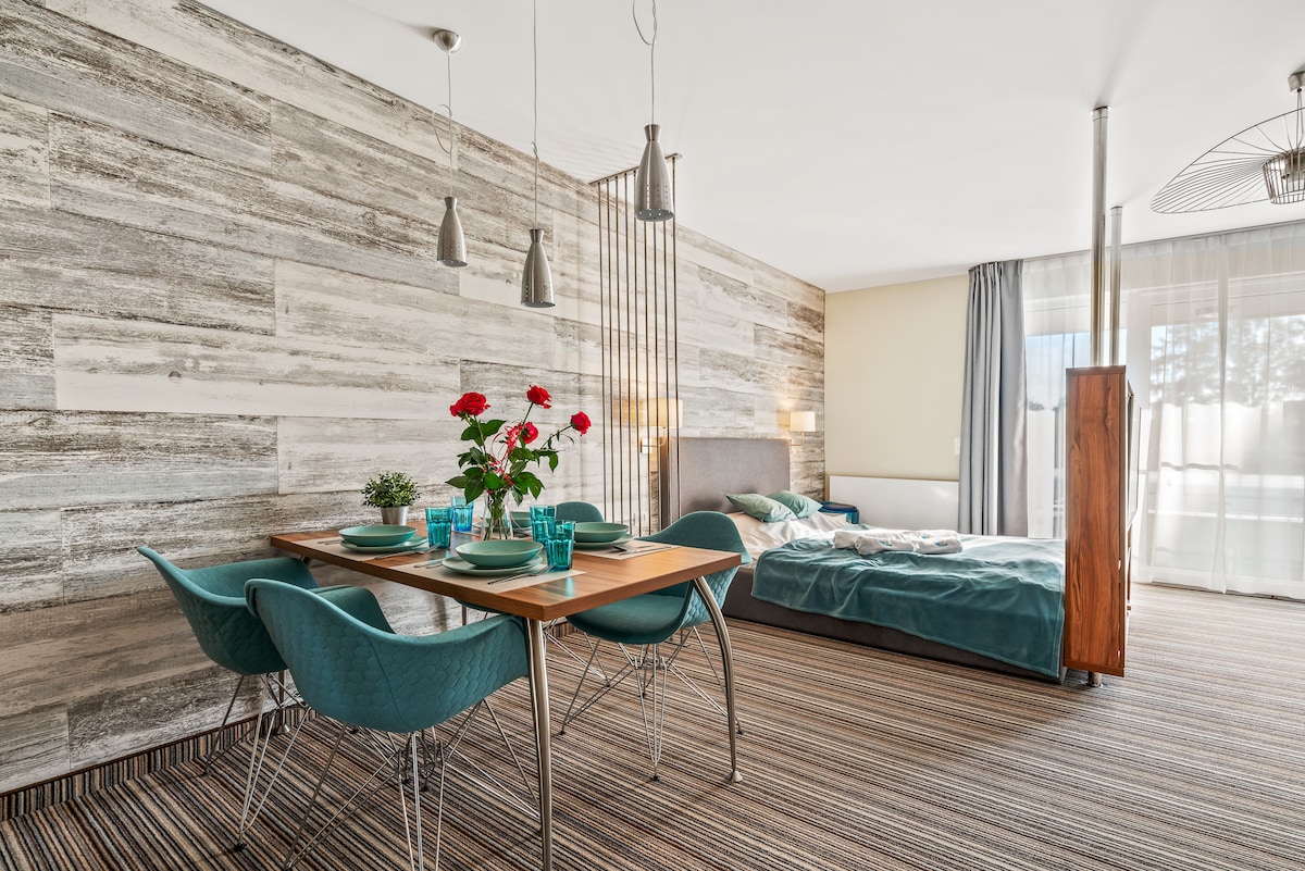 A bright living space features a dining area with a wooden table set for four, complemented by turquoise chairs. A spacious bed with soft bedding is positioned nearby, and large windows allow natural light to brighten the room, enhancing the wood-paneled accent wall.