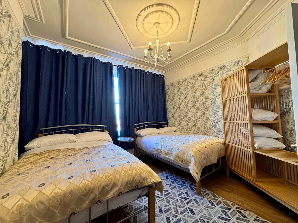 A bedroom featuring two spacious beds dressed in neutral linens. The walls are adorned with a subtle floral pattern. A stylish chandelier hangs from the ceiling, and large dark curtains frame a window, creating a cozy ambiance. A bamboo wardrobe provides storage and hanging space.