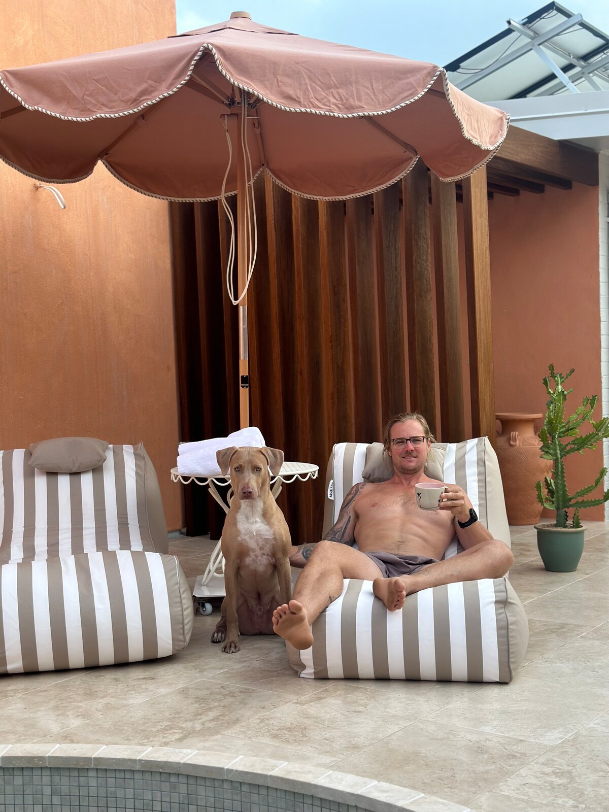 Two lounge chairs with striped fabric are positioned beside a magnesium plunge pool. A person is seated on one chair with a beverage, while a dog is calmly sitting beside them. An umbrella provides shade, and a small table is placed nearby.