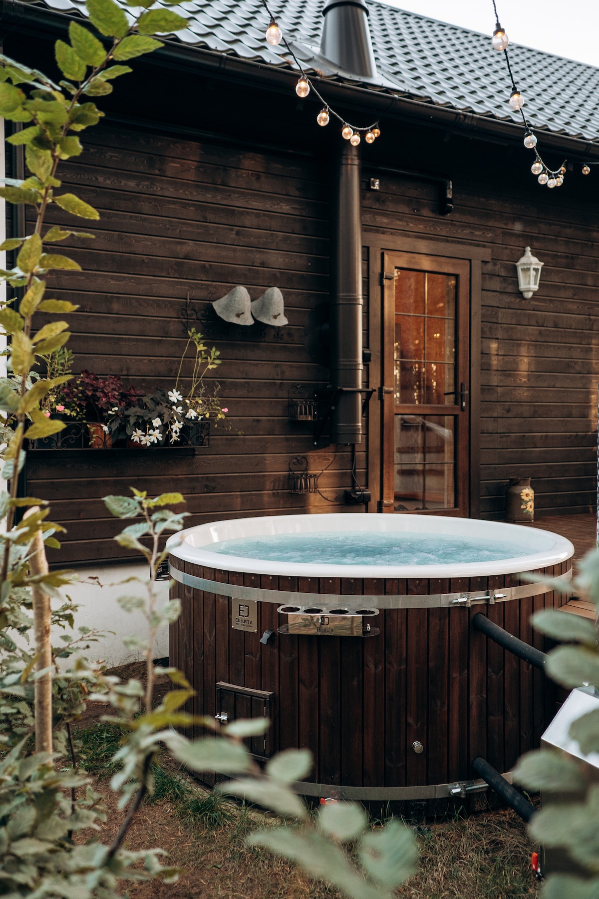 A wooden hot tub is situated in a garden area, complemented by greenery and soft outdoor lighting. The structure features a large, circular design and is positioned near a cozy wooden building with a welcoming entry door.
