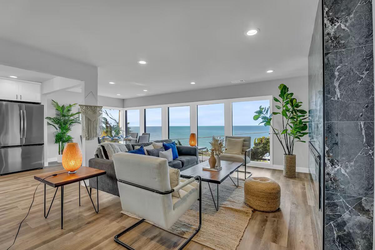 A spacious living area is showcased with large windows offering expansive ocean views. The room features a modern sofa, a sleek dining table, and a textured area rug. Green plants add a touch of nature, while a warm light creates a cozy ambiance.