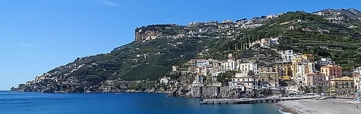 The image captures a coastal view of a hillside town along the Amalfi Coast, featuring colorful buildings nestled against a green mountain backdrop. A sandy beach stretches along the shoreline, with gentle waves lapping at the shore and a clear blue sky overhead.