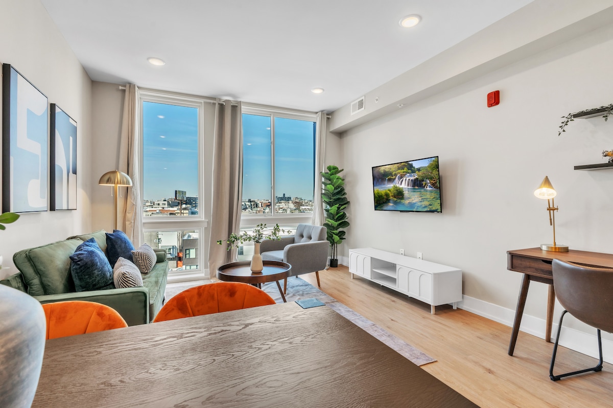 A bright living area features large windows offering urban views. Comfortable seating is arranged, including a cozy sofa with colorful cushions. A wall-mounted TV is positioned opposite a stylish coffee table, while a workspace is present with a small desk and chair, creating a functional environment.