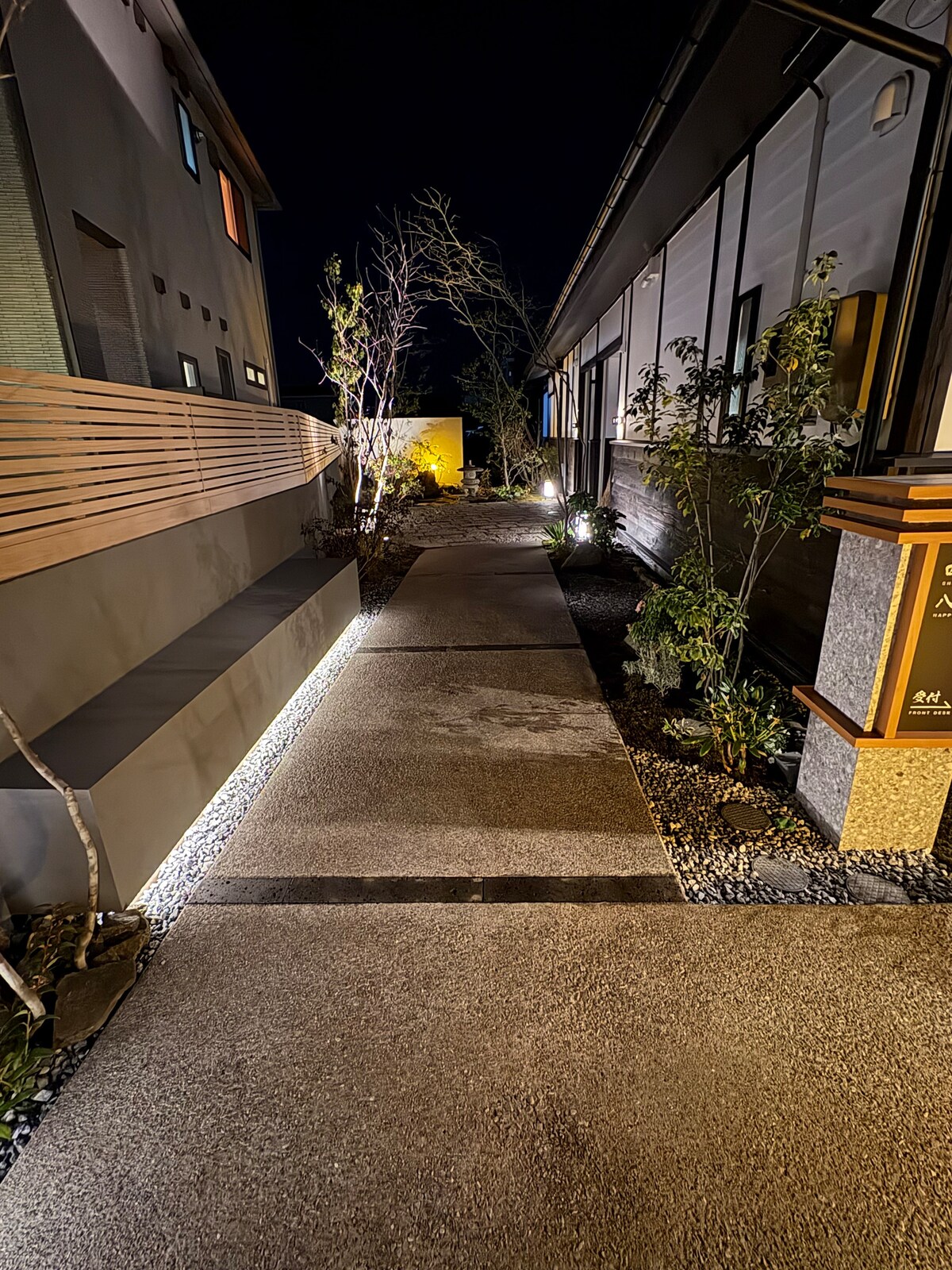 A well-lit pathway is seen between two buildings, featuring smooth stone surfaces bordered by landscaped greenery. Soft lighting enhances the path's atmosphere, leading the way to an inviting entrance. Trees and shrubs are strategically placed along the sides, contributing to a serene environment.