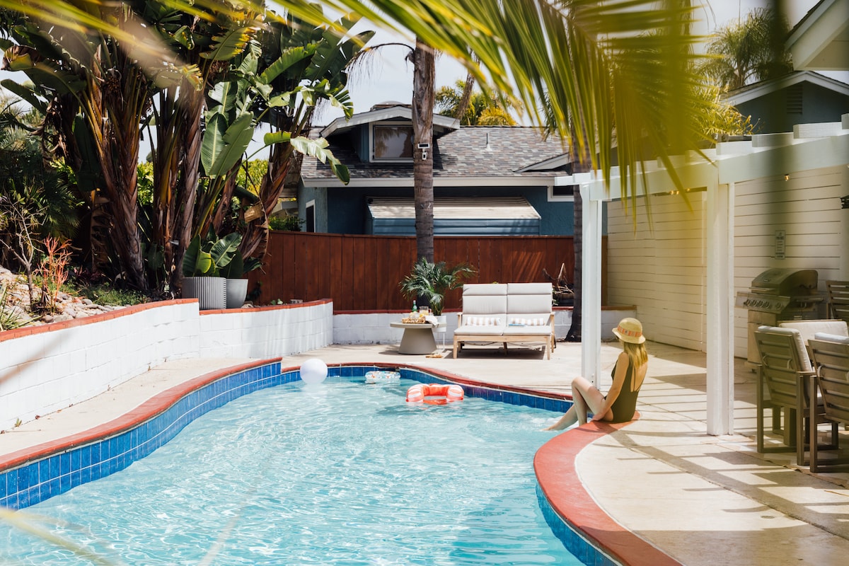 A private pool is surrounded by palm trees and a spacious patio area. Comfortable lounge chairs and a small table are positioned near the water. The clear blue water invites relaxation, and colorful pool floats are visible in the pool.