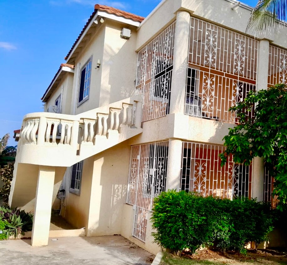 A two-story guest house features a decorative railing leading up to a second-floor entrance. The exterior is light-colored with large windows, some adorned with intricate metal grilles, and surrounded by lush greenery and well-maintained landscaping.