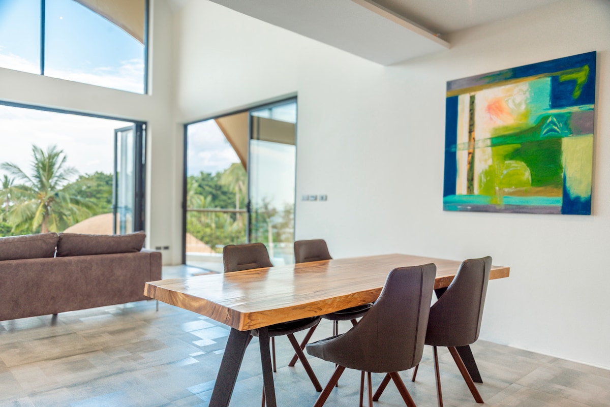 A spacious dining area features a large wooden table surrounded by four modern chairs. Large glass doors invite natural light and showcase views of the lush outdoor landscape, with palm trees in the background. A colorful artwork adorns the wall, adding visual interest.