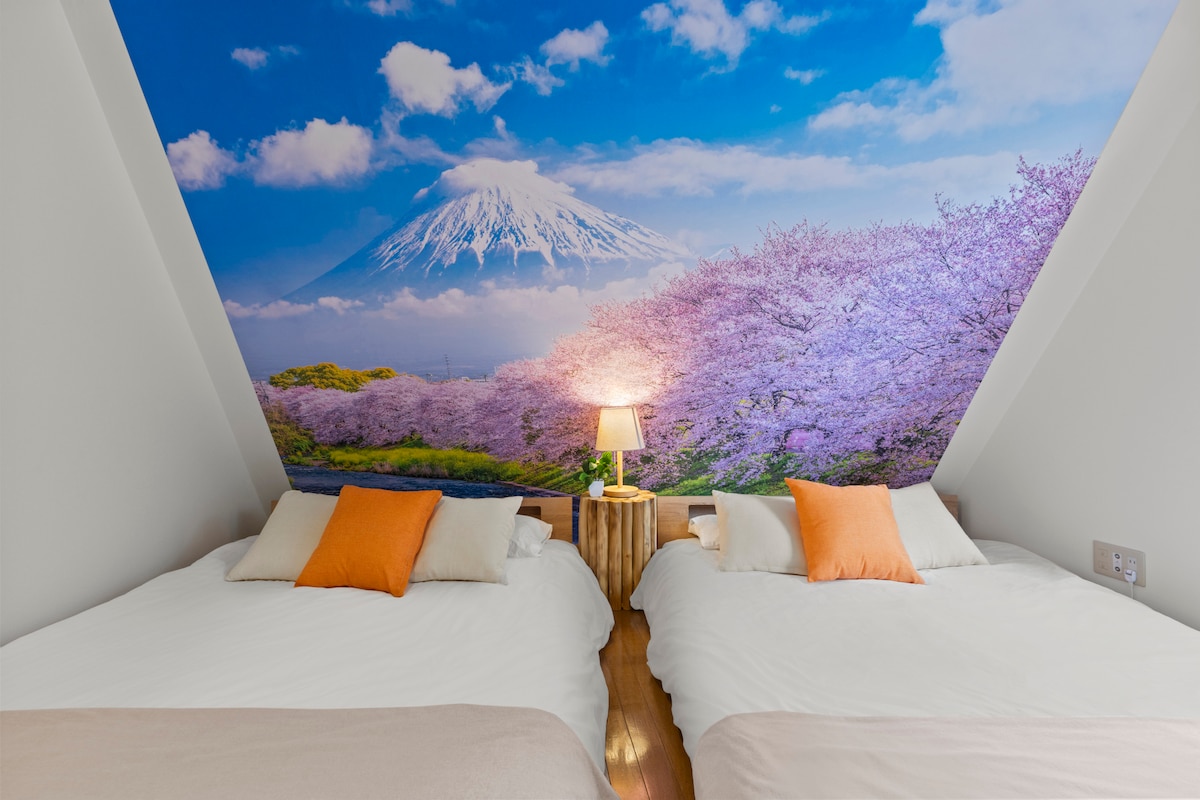 A serene sleeping area features two spacious beds with light bedding and vibrant orange accent pillows. Behind the beds, a striking mural depicts Mount Fuji surrounded by blooming cherry blossoms under a bright sky. A small lamp rests on a wooden nightstand, adding a soft glow.