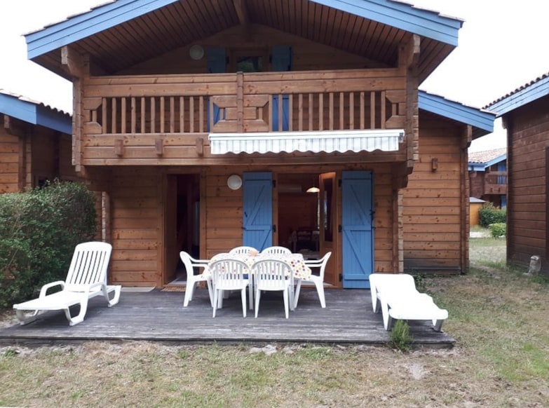 A wooden chalet features a spacious outdoor terrace with a large table surrounded by several white chairs. Two lounge chairs can be seen, providing a relaxing space. The building stands in a natural setting, with wooden architecture and blue accents complementing the tranquil environment.