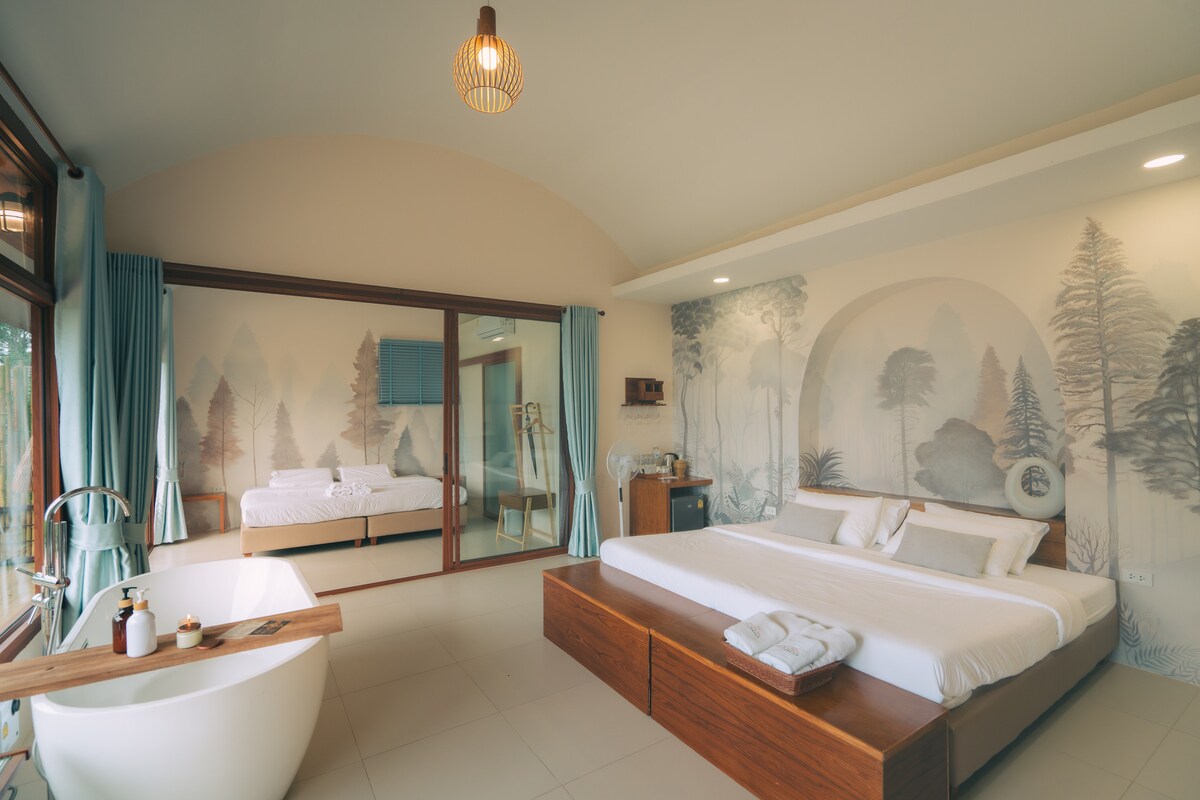 A spacious bedroom is illuminated by natural light, featuring a large bed with neutral-toned linens and soft pillows. A freestanding bathtub is positioned nearby, alongside a wooden table. The walls are adorned with serene tree-themed murals, creating a calming ambiance.