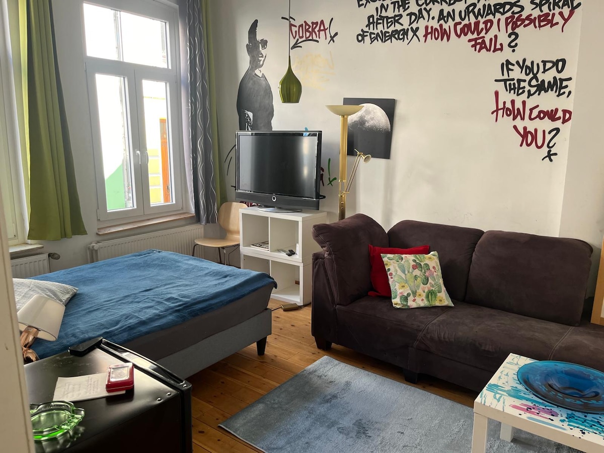 A living area features a spacious bed with a blue coverlet, accompanied by a dark sofa adorned with a decorative pillow. A small television is positioned on a white stand, while a light-green lamp and a wooden chair add to the decor. Bright artwork and green curtains enhance the space.