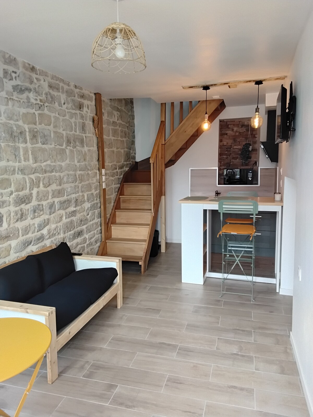 The living area features a light-colored sofa with a contrasting table, adjacent to a modern kitchen with bar seating for four. A staircase leads to the upper level, with stone walls providing a rustic touch. Pendant lights hang from the ceiling, enhancing the space.