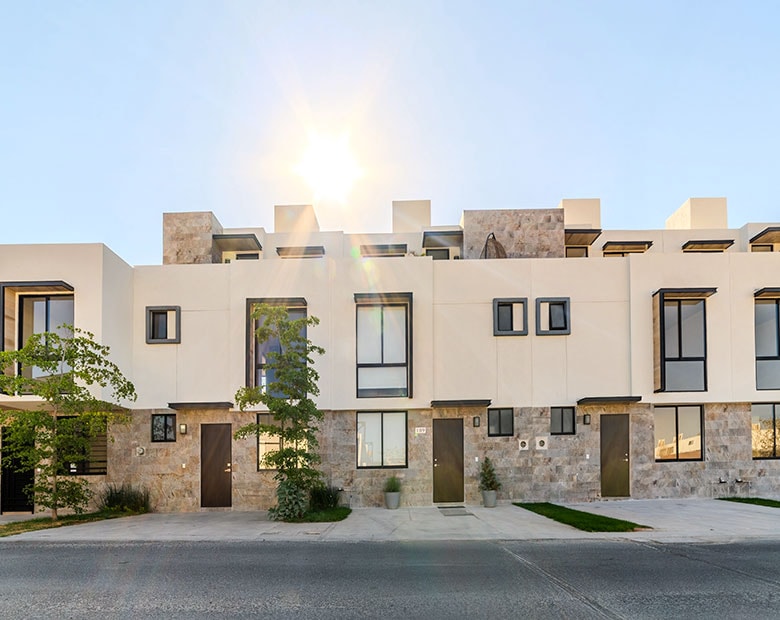 A modern townhouse is showcased with a sleek exterior featuring a mix of stone and smooth white surfaces. Sunlight reflects off the building, creating a warm ambiance. Large windows and a minimalist design enhance the inviting appearance, complemented by landscaping that adds a touch of greenery.