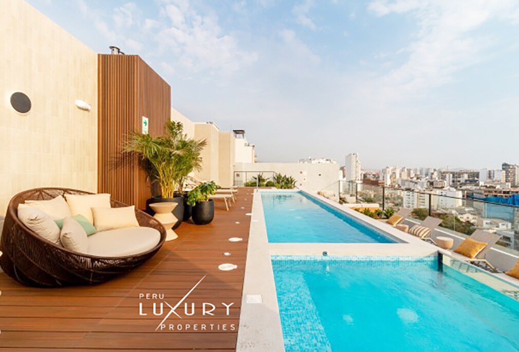 A rooftop terrace features a swimming pool with a clear blue surface and surrounding wooden decking. Comfy lounge seating is positioned next to lush greenery, offering scenic views of the city skyline in the background under a partially cloudy sky.