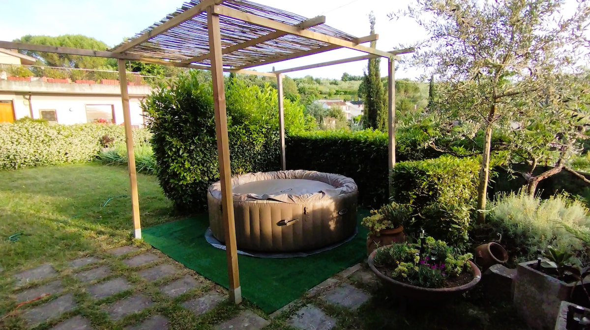 A private outdoor area features a circular, inflatable hot tub situated on a grassy patch. Surrounding greenery offers natural privacy, and a wooden pergola provides partial shade. A pathway made of stone pavers leads to the hot tub, contributing to the serene garden atmosphere.