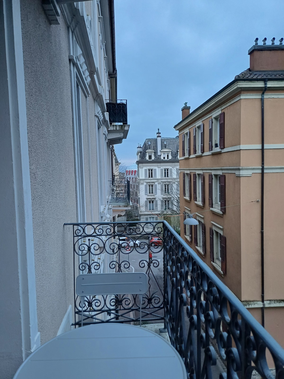 A narrow balcony features a small round table surrounded by elegant wrought iron railings. The view showcases nearby buildings, with architectural details visible in pastel shades. A hint of the street below is seen, revealing a quiet urban setting under an overcast sky.